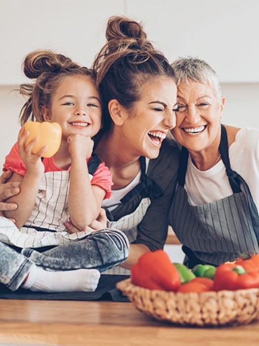 Three generations of women laughing and cooking in a kitchen.