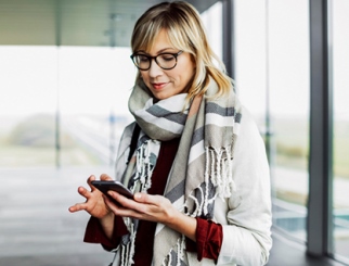 Blonde woman wearing a sweater, scarf and glasses and looking down at her cell phone.